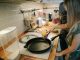 Young girl cooking with a frying pan under adult supervision in a modern kitchen.